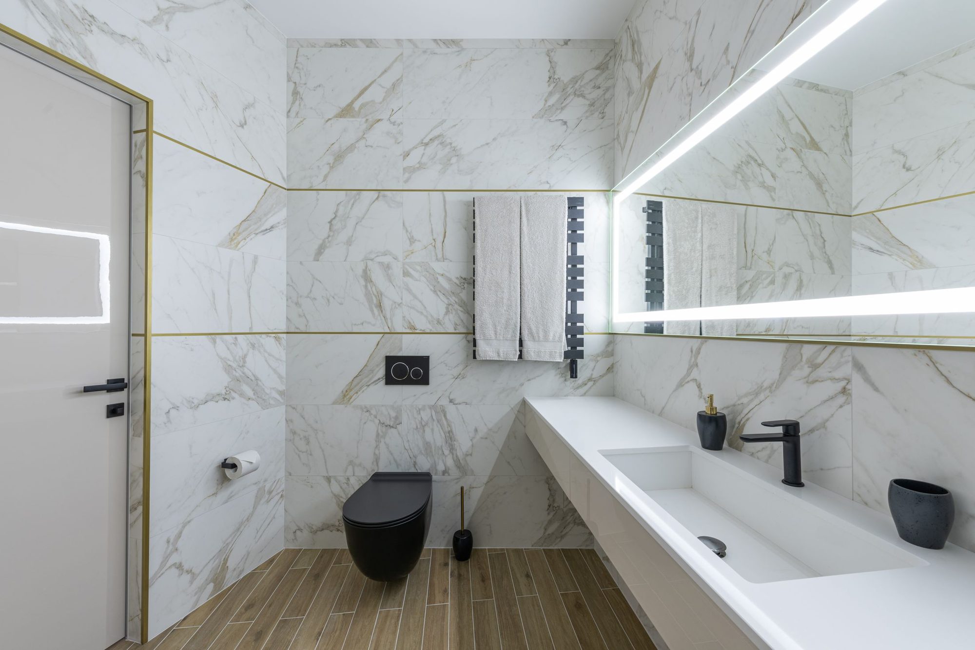 A marble-clad bathroom with a slim brass keyline set into the wall, a back-lit mirror and a wall-hung WC