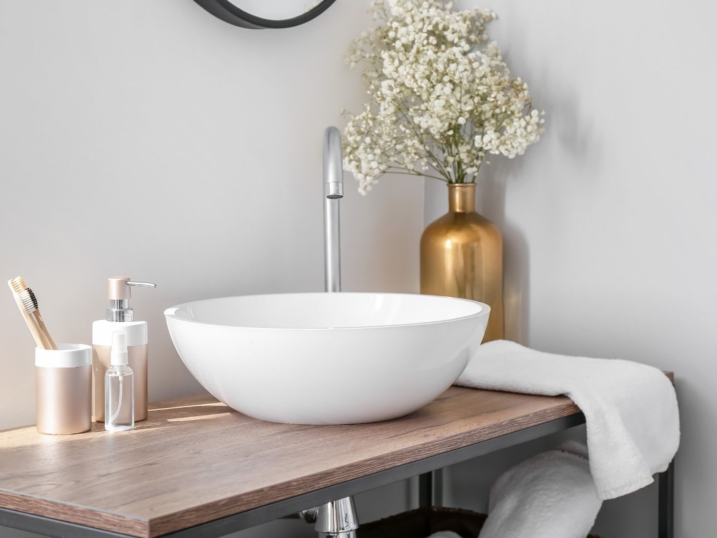 A white countertop basin on an oak vanity with a chrome tap, folded towel and a brass vase