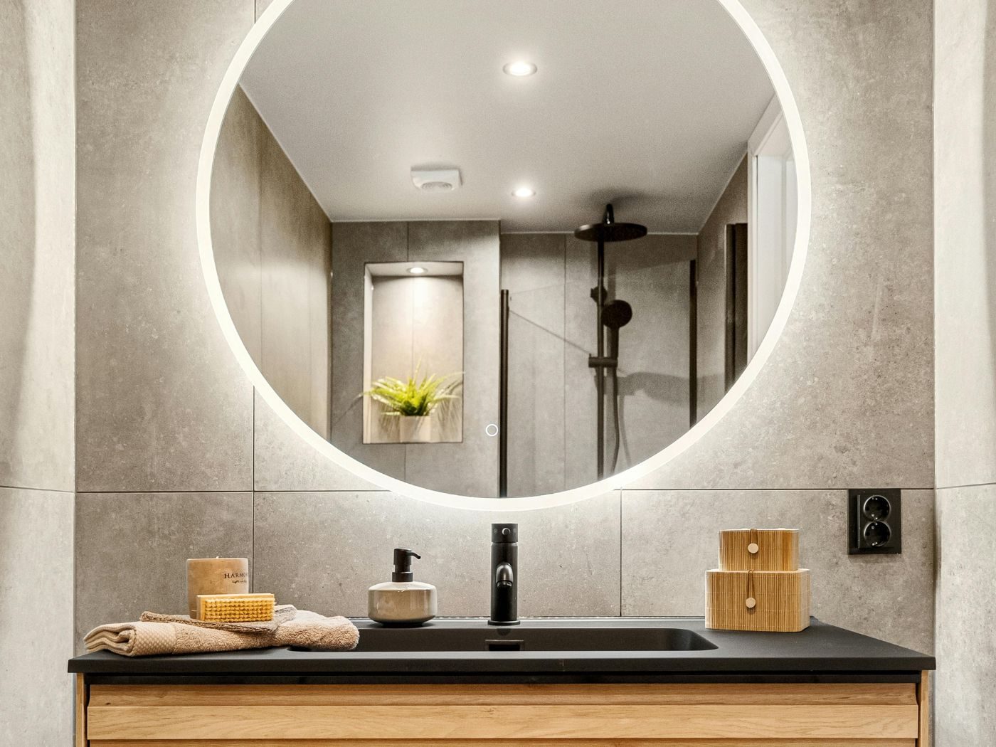 A bathroom tiled in large concrete-effect porcelain with a circular back-lit mirror over a dark basin